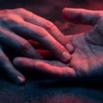 Amor-en-linea.com mon homme m'aime t'il : 10 Signes Secrets 20 Gros plan sur deux mains qui se frôlent délicatement avec des reflets néon rouges et violets sur fond sombre.