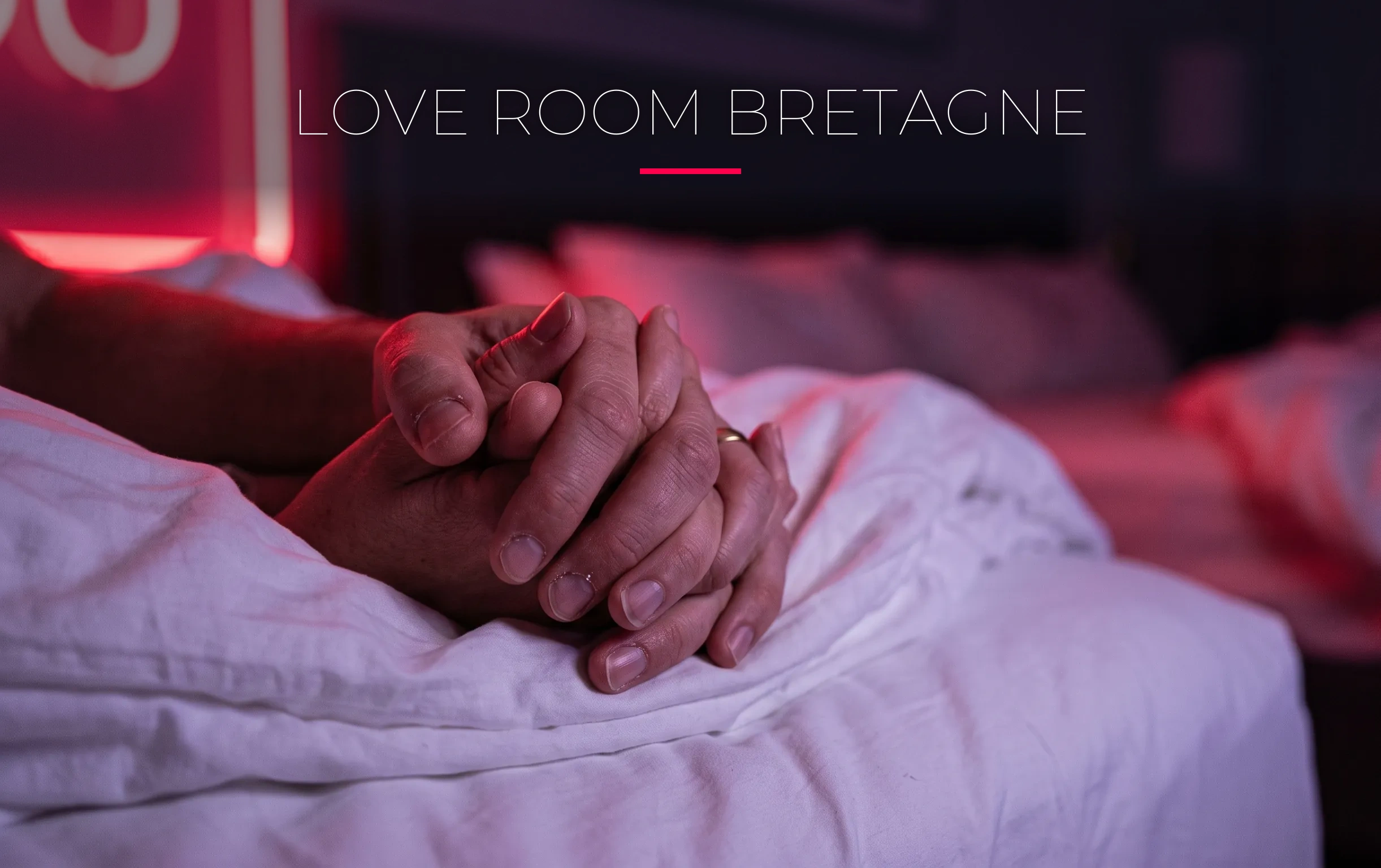Love room Bretagne : l'expérience vaut-elle le coup ? 1 Des doigts entrelacés sur des draps blancs dans une love room bretonne éclairée par une lumière néon rouge cerise