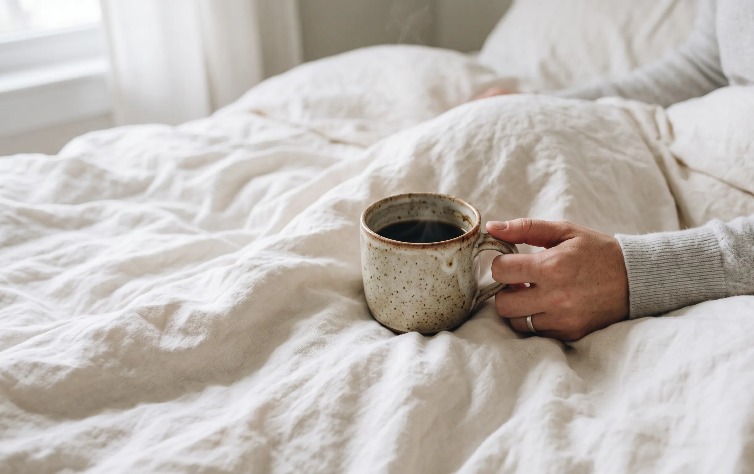 comment avouer ses sentiments à son crush : Une main tient une tasse en céramique sur des draps blancs froissés dans la lumière froide du matin